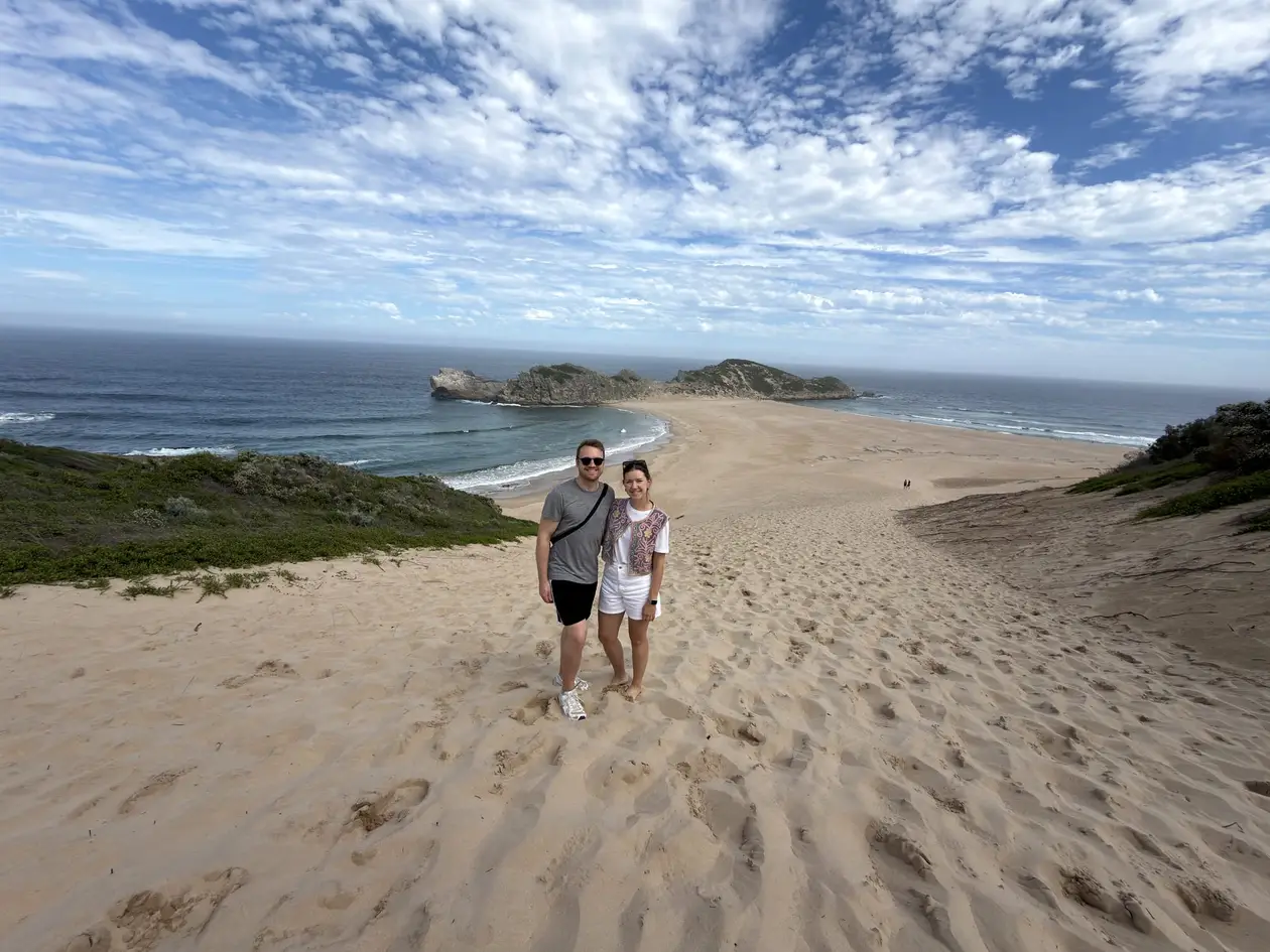 Max und Rebecca stehen zusammen im Sand am Wasser, im Hintergrund eine Felsinsel