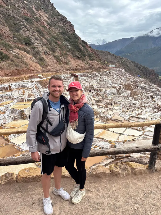 Max und Rebecca stehen an einem Holzgeländer, hinter ihnen die terrassierten Salzbecken von Maras im Tal