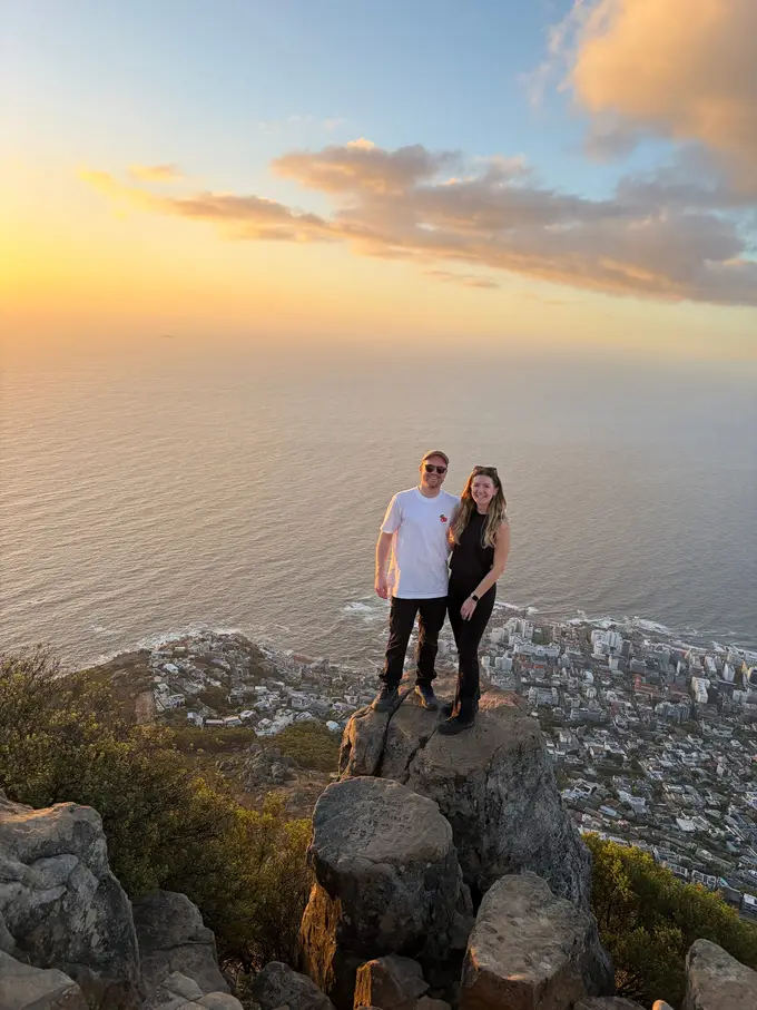 Max und Rebecca stehen zusammen auf einem Felsen oben auf dem Lion's Head, dahinter das Meer im Sonnenuntergang