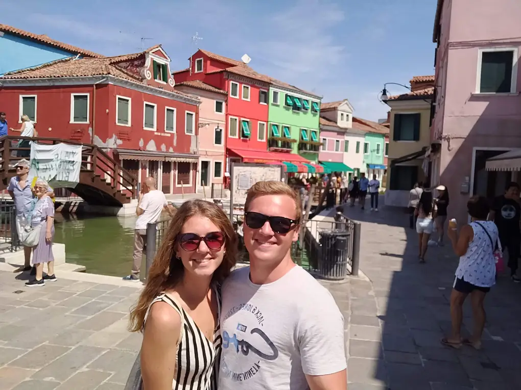 Rebecca und Max vor roten, grünen und rosa Häusern an einem Kanal mit einer Holzbrücke, ringsum Spaziergänger