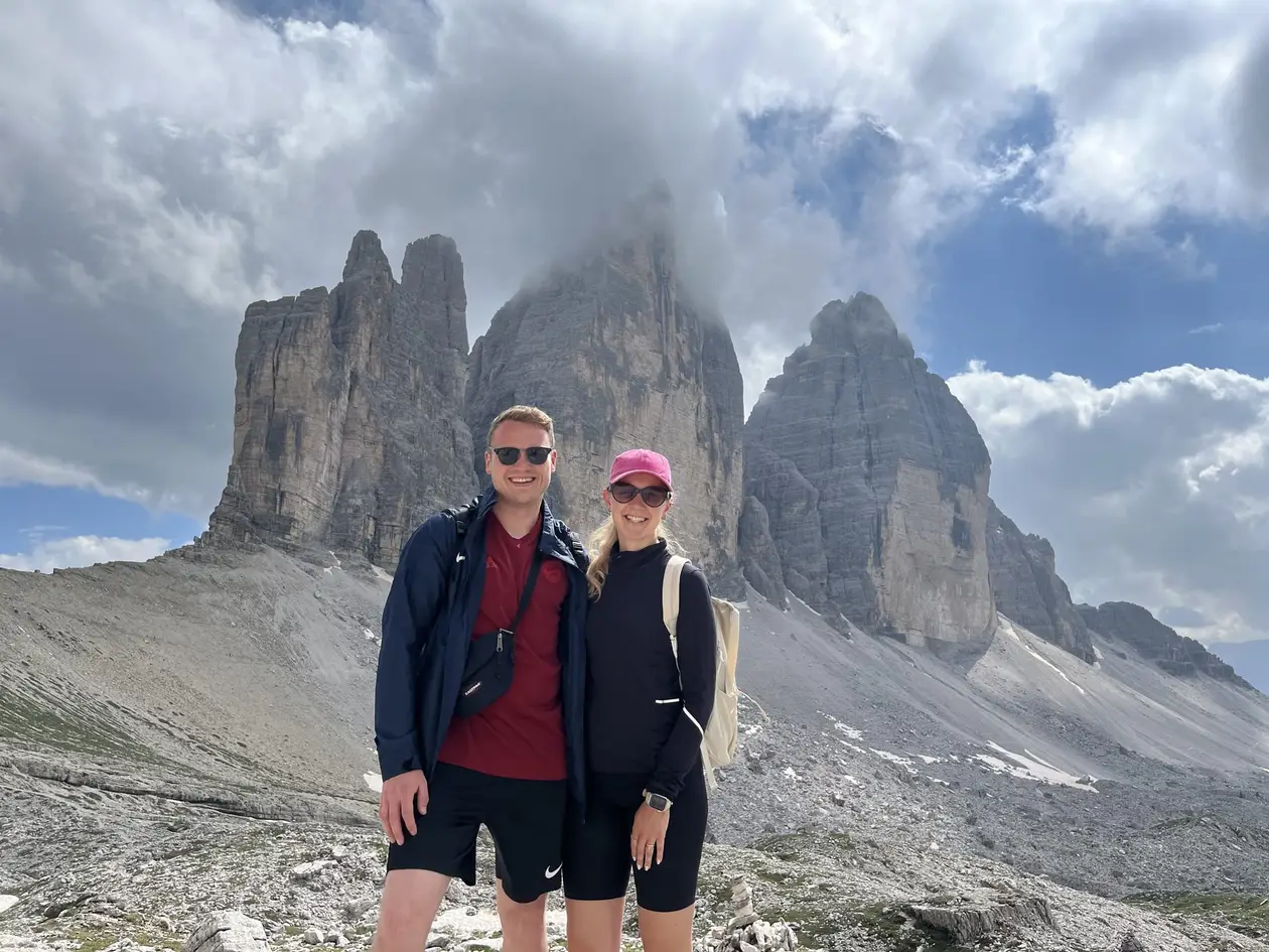 Max und Rebecca stehen lächelnd auf steinigem Boden, hinter ihnen die drei Felstürme der Drei Zinnen, die Spitze des mittleren Turms in einer Wolke