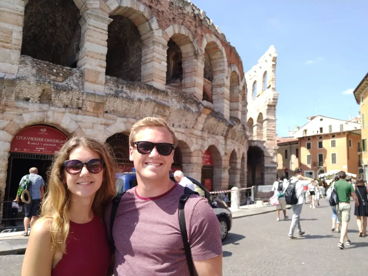 Rebecca und Max mit Sonnenbrillen vor den Bögen der römischen Arena, rechts bunte Häuser und Passanten