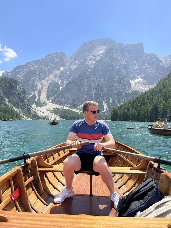 Max sitzt mit beiden Rudern in der Hand in einem hölzernen Ruderboot, hinter ihm der See, weitere Boote und das Felsmassiv