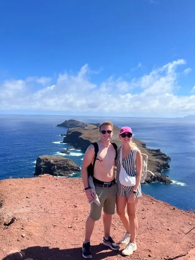 Max und Rebecca stehen lächelnd mit Rucksack und Tasche auf einem Felsvorsprung, dahinter der Atlantik und die Ostspitze der Insel