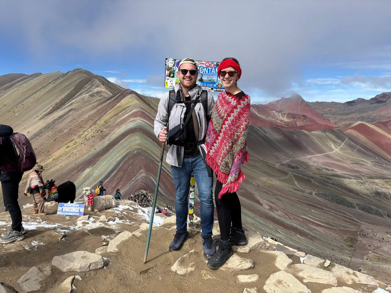 Max und Rebecca am Gipfelschild, links die gestreiften Hänge des Rainbow Mountain, rechts rote Berge