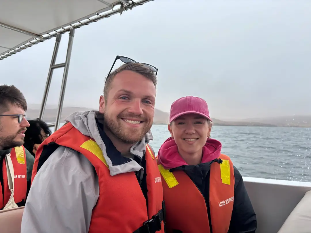 Max und Rebecca in orangen Schwimmwesten sitzen lachend im Boot, hinter ihnen graues Meer und Küste im Dunst
