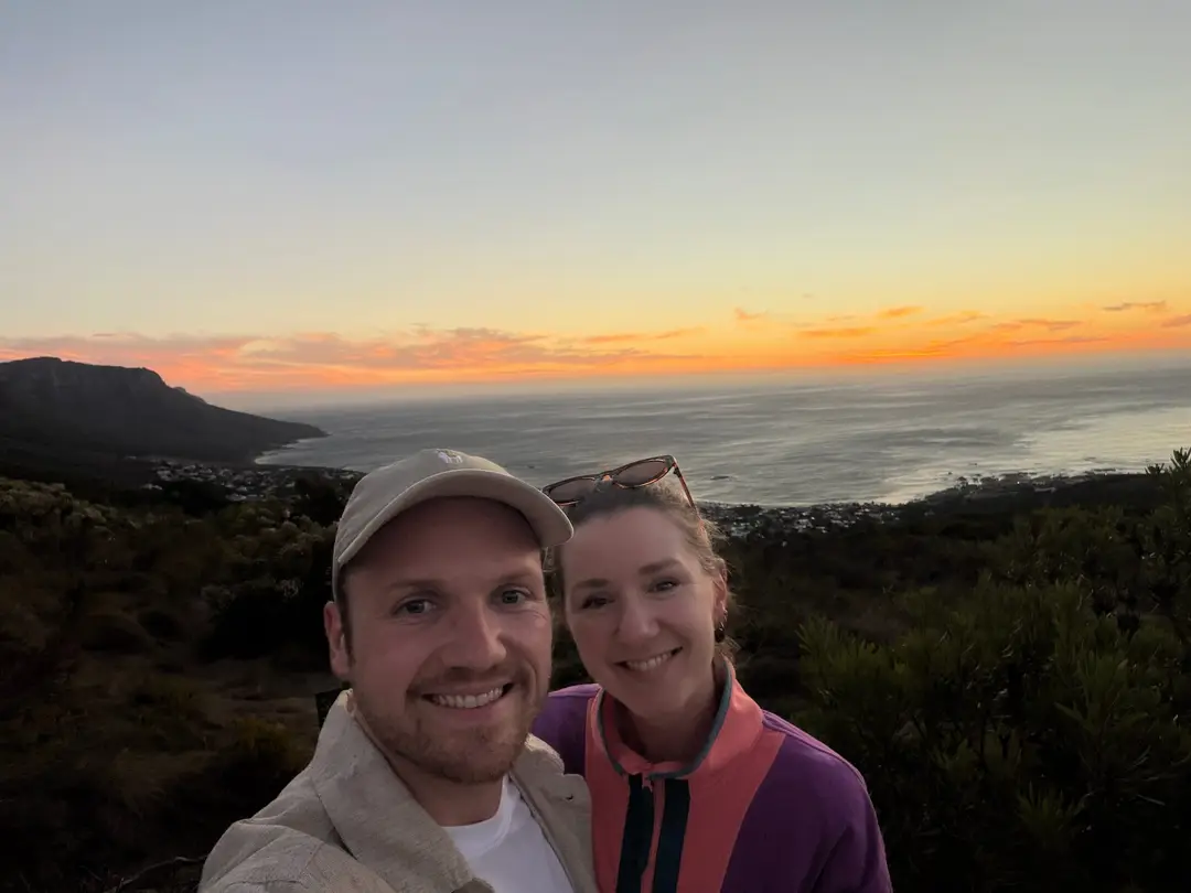 Max und Rebecca lächeln in die Kamera, dahinter ein orangefarbener Sonnenuntergang über der Küste