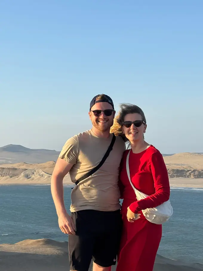 Max und Rebecca stehen mit Sonnenbrillen auf einer Anhöhe, hinter ihnen eine Meeresbucht und helle Wüstenhügel