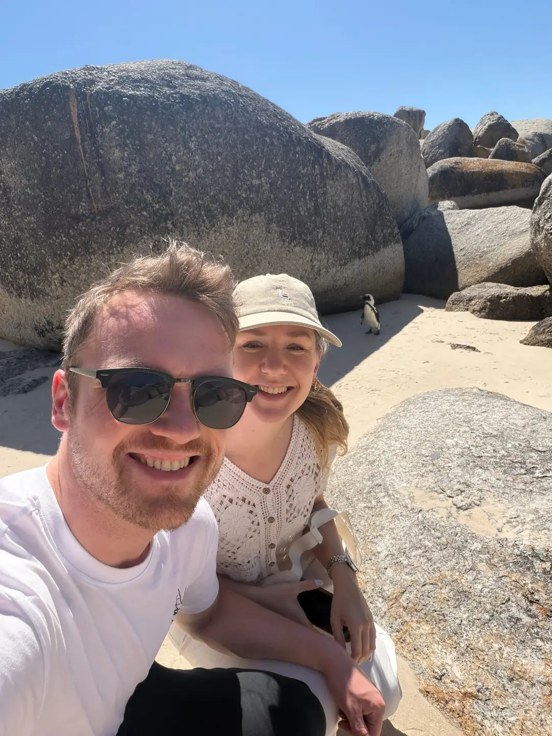 Max und Rebecca lächeln in die Kamera, im Hintergrund ein Pinguin zwischen großen Granitfelsen