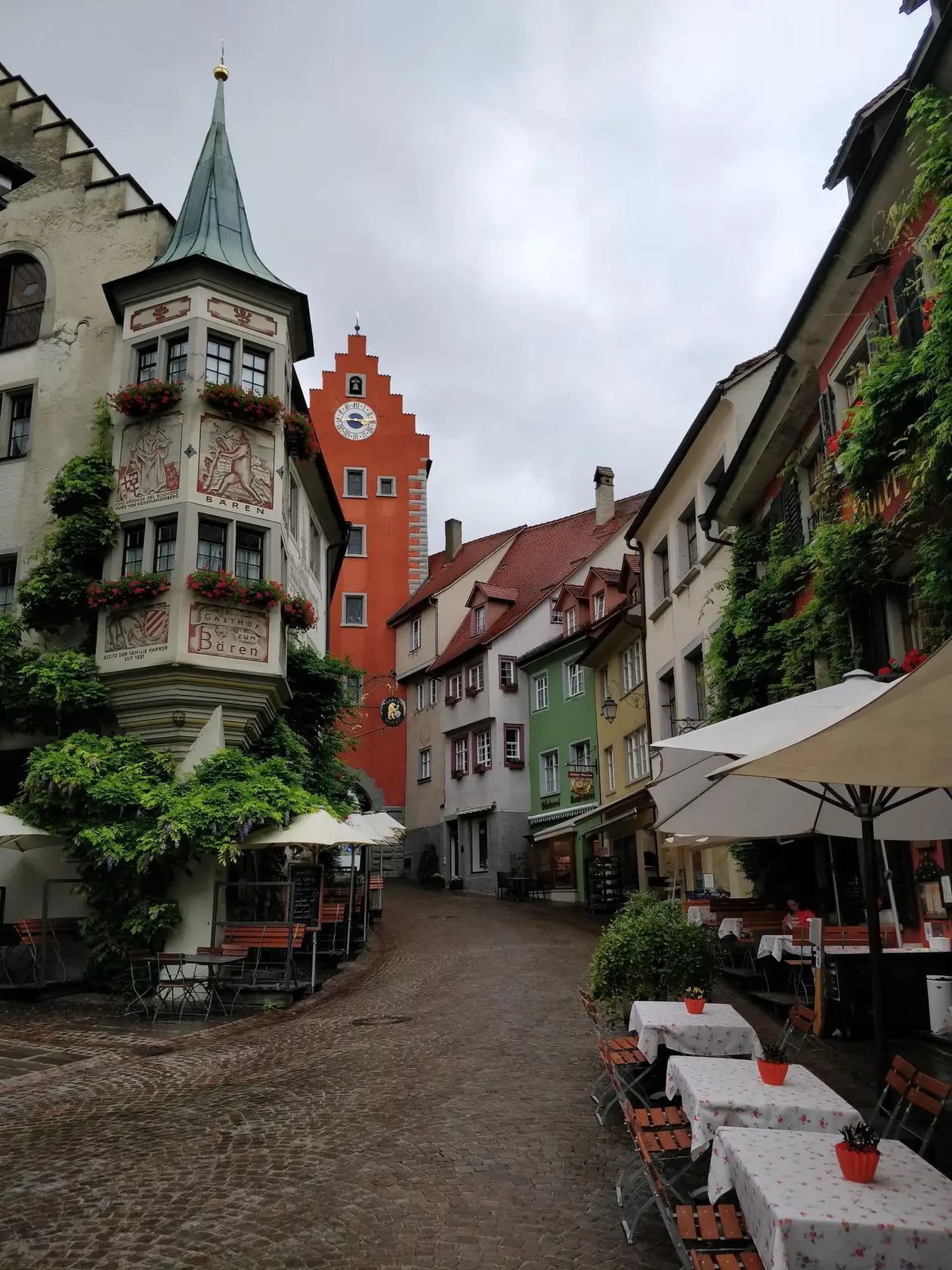 Gepflasterte Altstadtgasse mit Tischen und Sonnenschirmen, links ein bemalter Erker mit grünem Spitzdach, in der Mitte ein rotes Tor mit Treppengiebel und Uhr