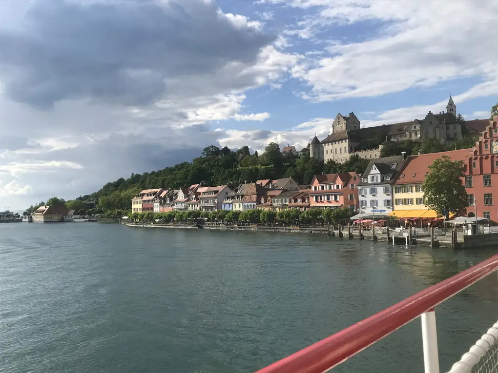 Meersburg vom Schiff aus, eine Reihe bunter Häuser an der Uferpromenade, darüber auf dem Hang die Burg, vorne die rote Reling