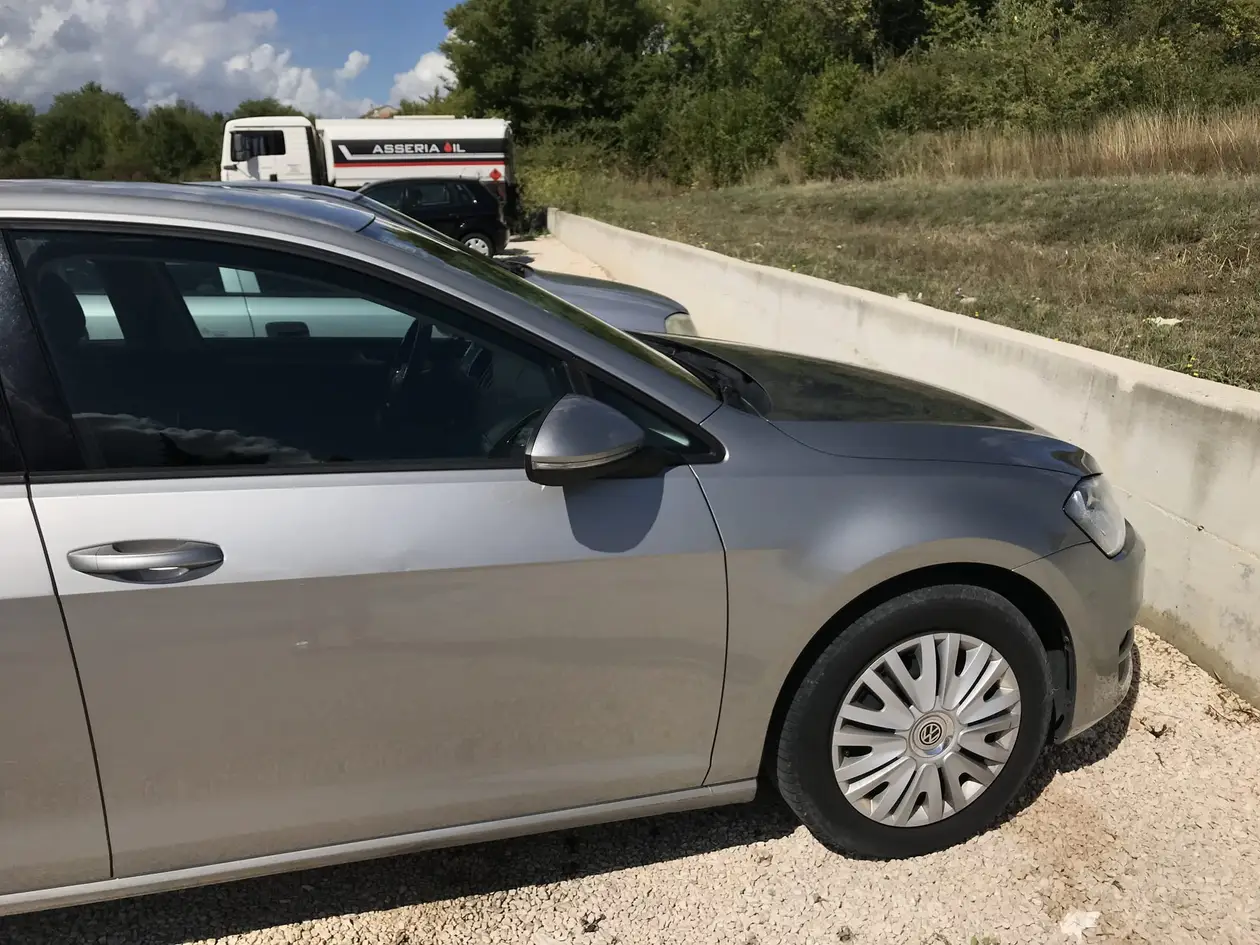 Ein silberner VW Golf auf einem Schotterplatz am Straßenrand, dahinter eine Betonmauer und ein geparkter Tankwagen