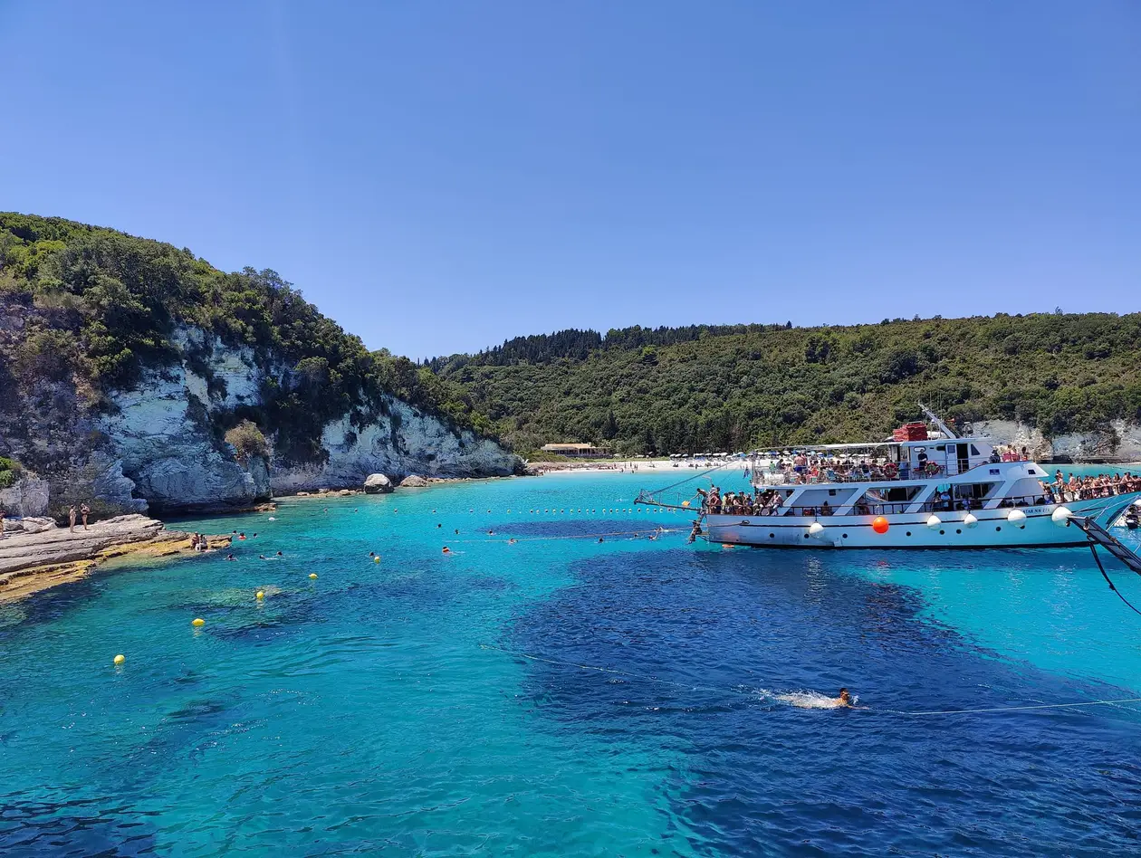 Leuchtend türkises Wasser über hellem Grund vor einer weißen Felsküste, in der Bucht ein vollbesetztes Ausflugsboot