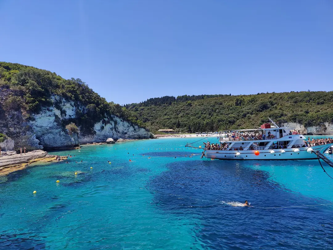 Leuchtend türkises Wasser über hellem Grund vor einer weißen Felsküste, in der Bucht ein vollbesetztes Ausflugsboot