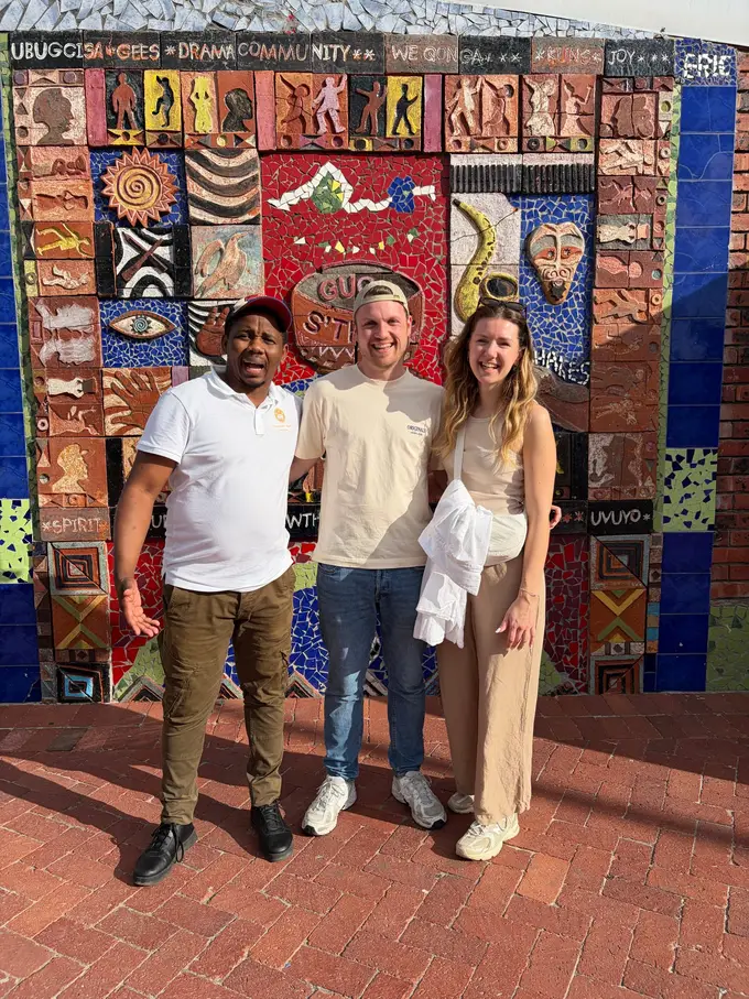 Der Guide steht zusammen mit Max und Rebecca vor der Mosaikwand des Kulturzentrums Guga S'thebe