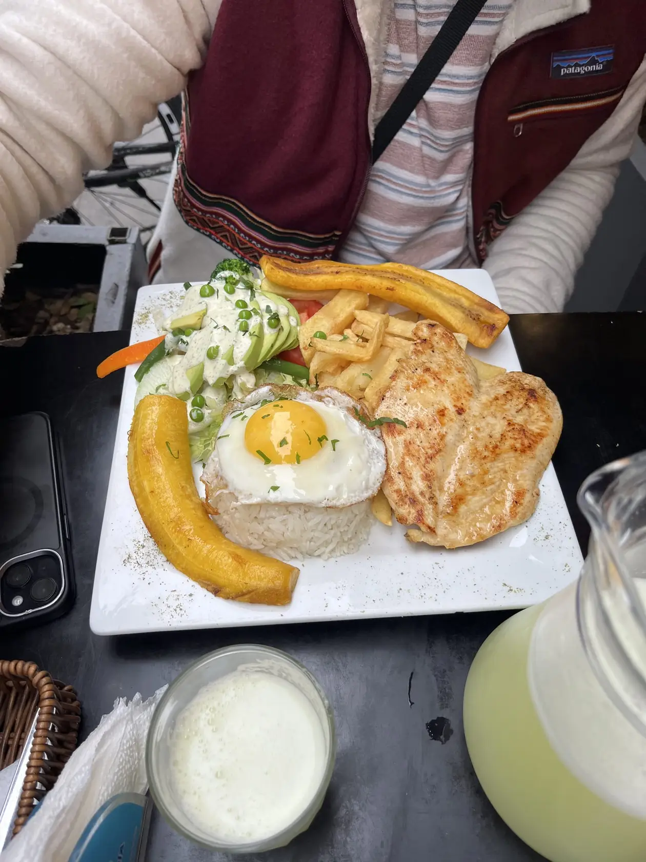 Teller mit gebratenem Hähnchenfilet, Spiegelei auf Reis, gebratener Kochbanane, Pommes und Salat, daneben ein Krug Limonade