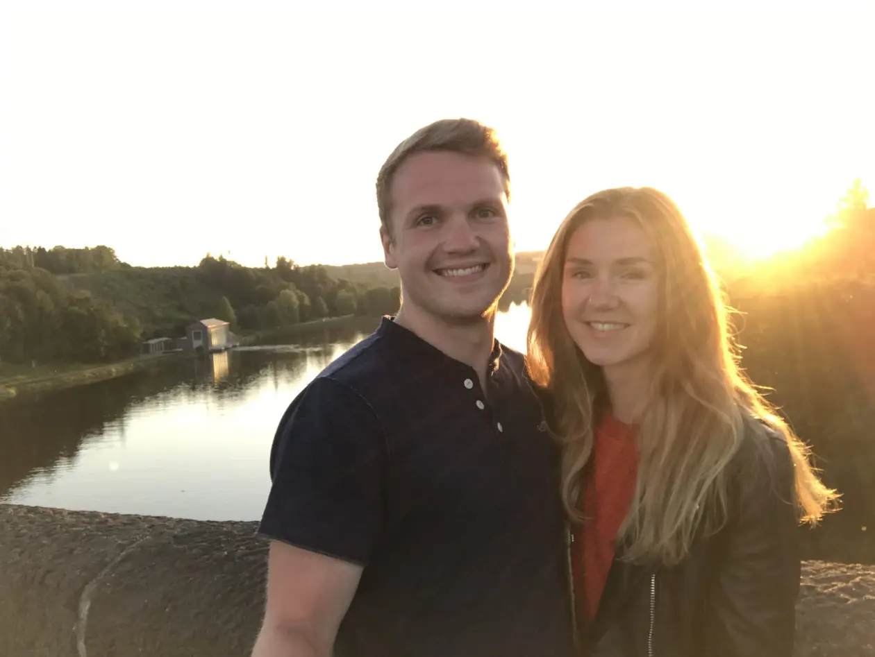 Max und Rebecca im Gegenlicht auf der Staumauer der Möhnetalsperre, hinter ihnen ein ruhiges Gewässer mit einem kleinen Gebäude am Ufer und rechts die tief stehende Sonne