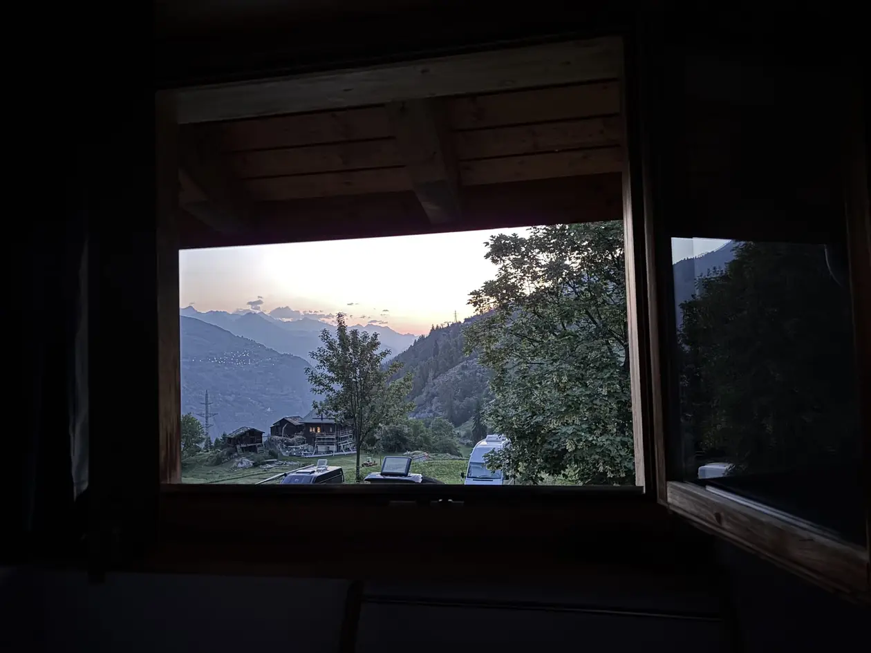 Durch ein offenes Fenster im dunklen Zimmer sieht man eine Wiese mit Holzhütte und Campingbussen, dahinter Berge in der Morgendämmerung