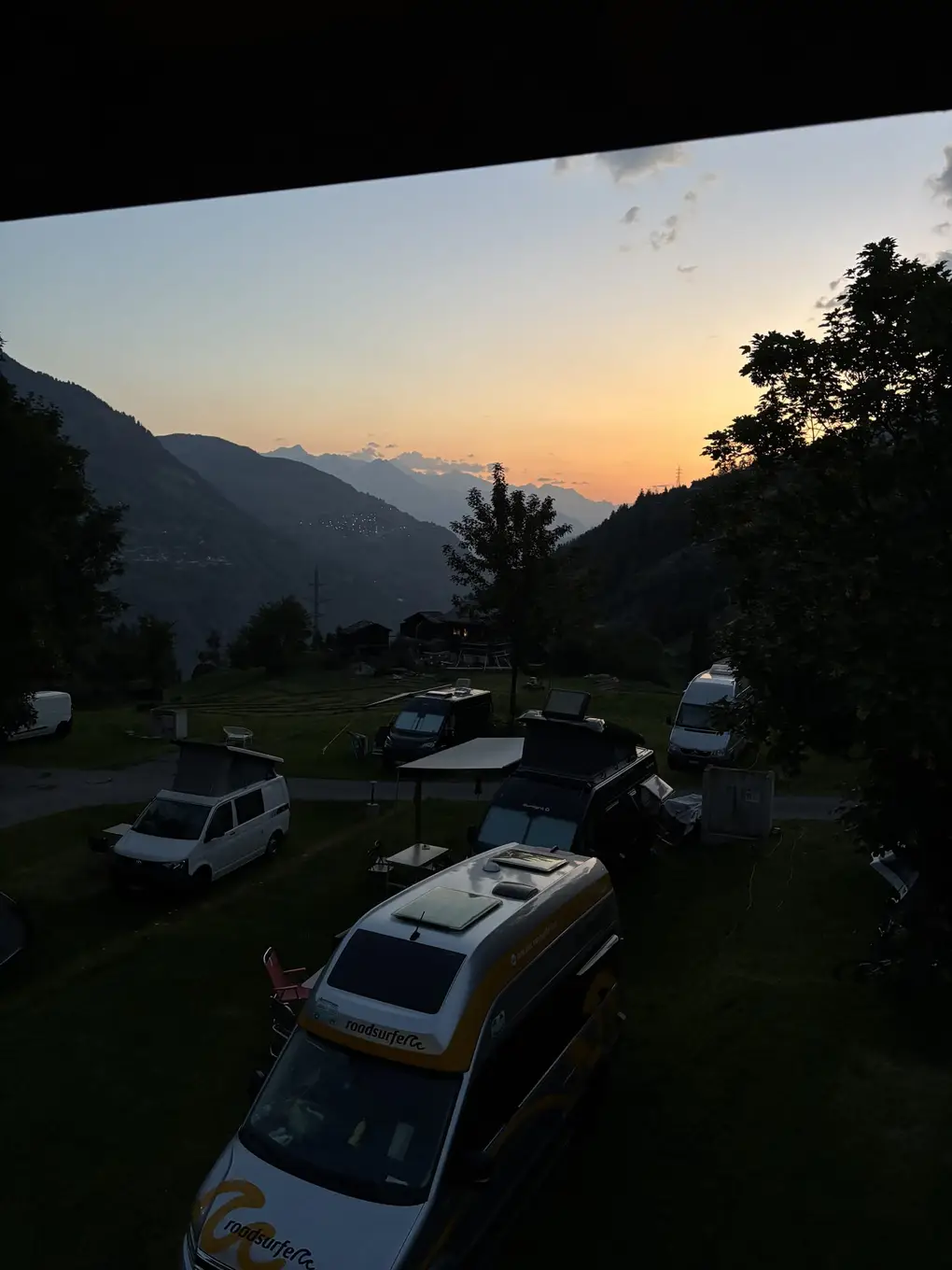 Morgenrot über einem tief eingeschnittenen Tal, im Vordergrund im Halbdunkel mehrere Campingbusse auf einer Wiese