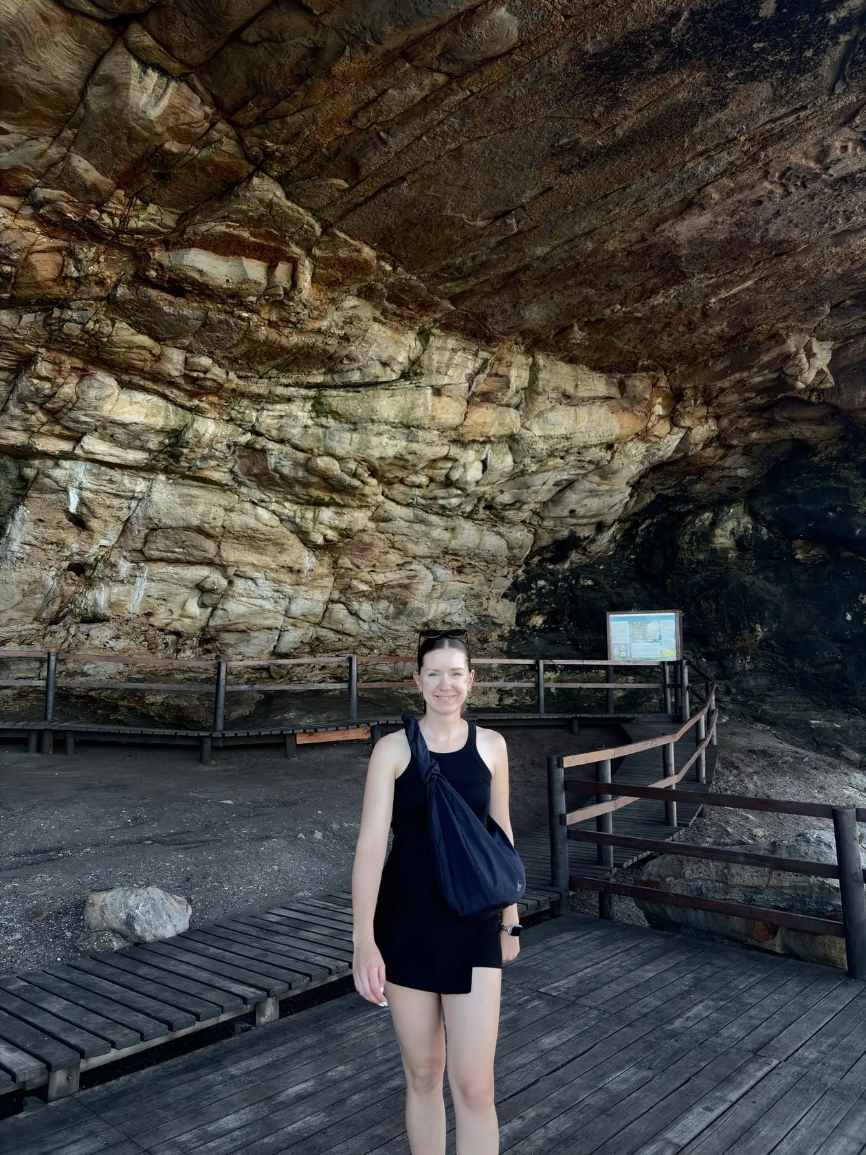 Rebecca steht auf einem Holzsteg vor einer großen Felshöhle an der Küste von Mossel Bay