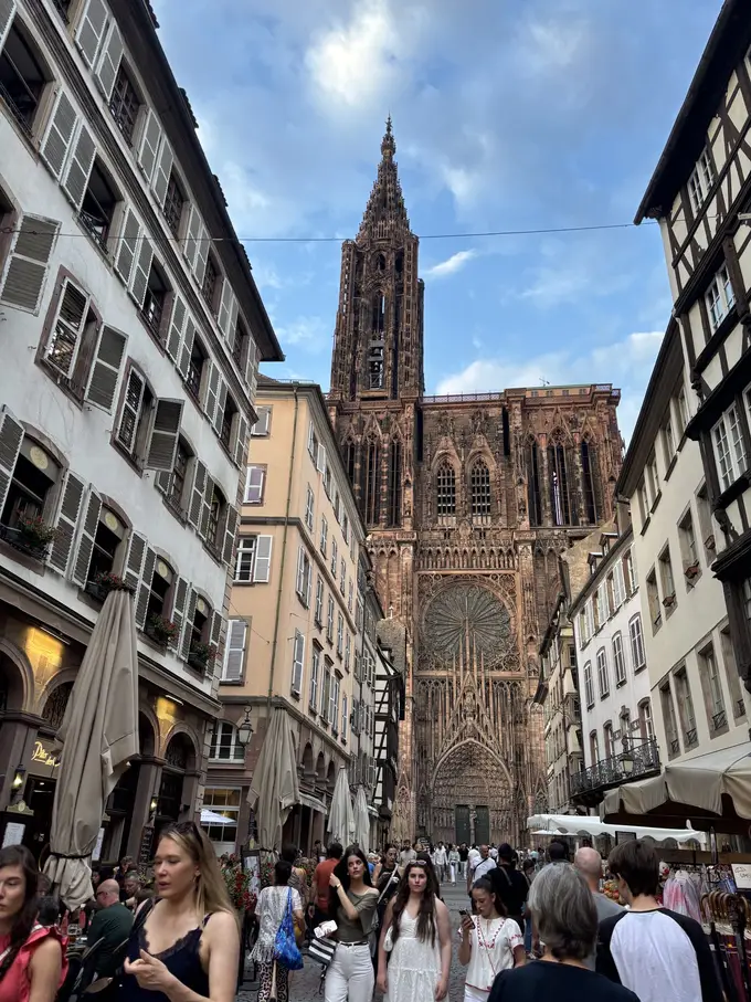 Eine volle Gasse mit hohen Häusern, an deren Ende die Westfassade des Münsters aus rotem Sandstein mit Rosette und Turm aufragt