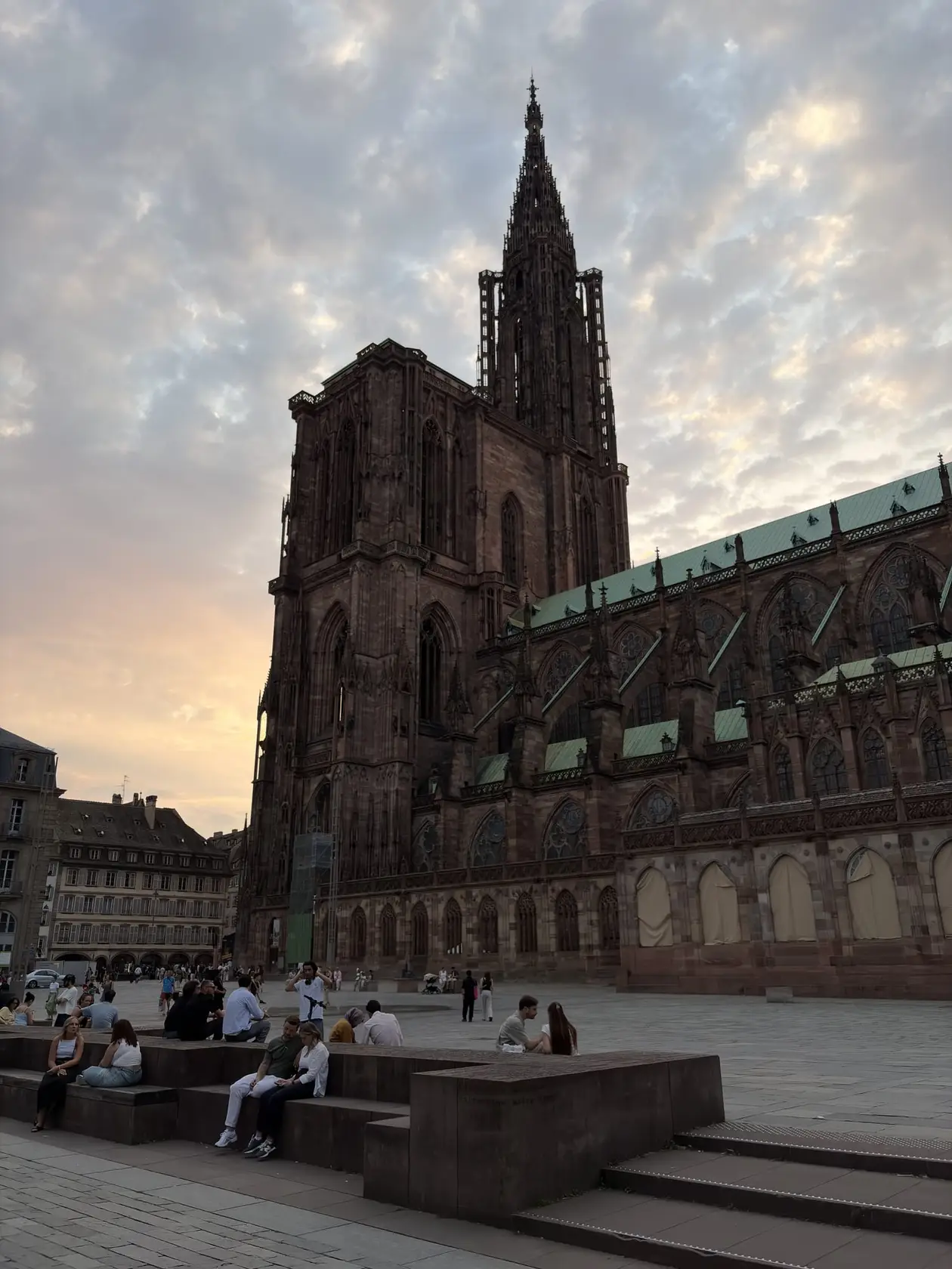 Das Straßburger Münster aus rotem Sandstein mit seinem einzelnen Turm vor einem Abendhimmel, davor Menschen auf einem Platz