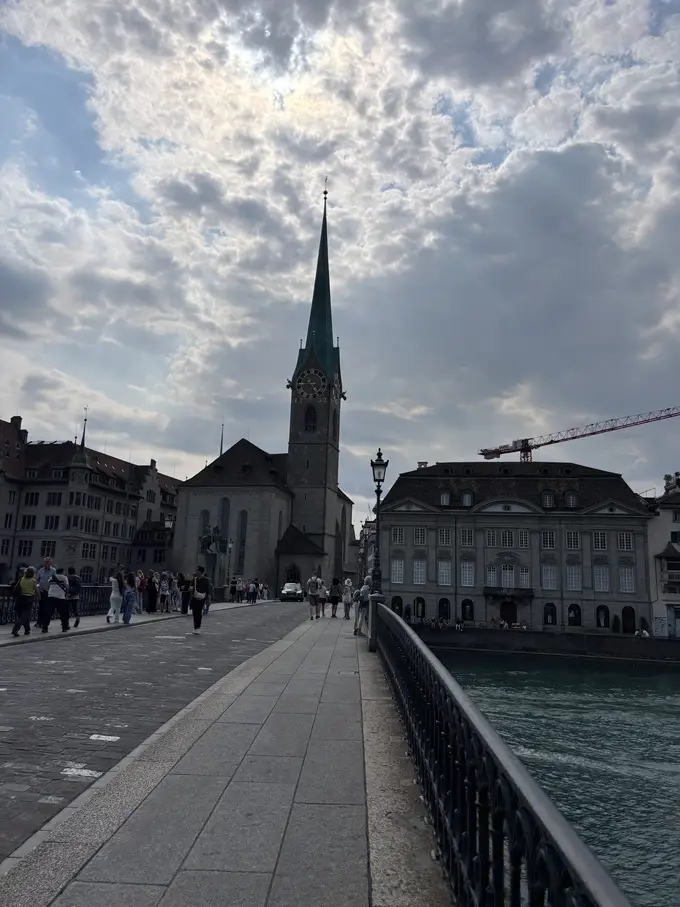 Auf einer Brücke über die Limmat, am Ende die Fraumünsterkirche mit spitzem grünem Turm, rechts ein barockes Gebäude am Wasser