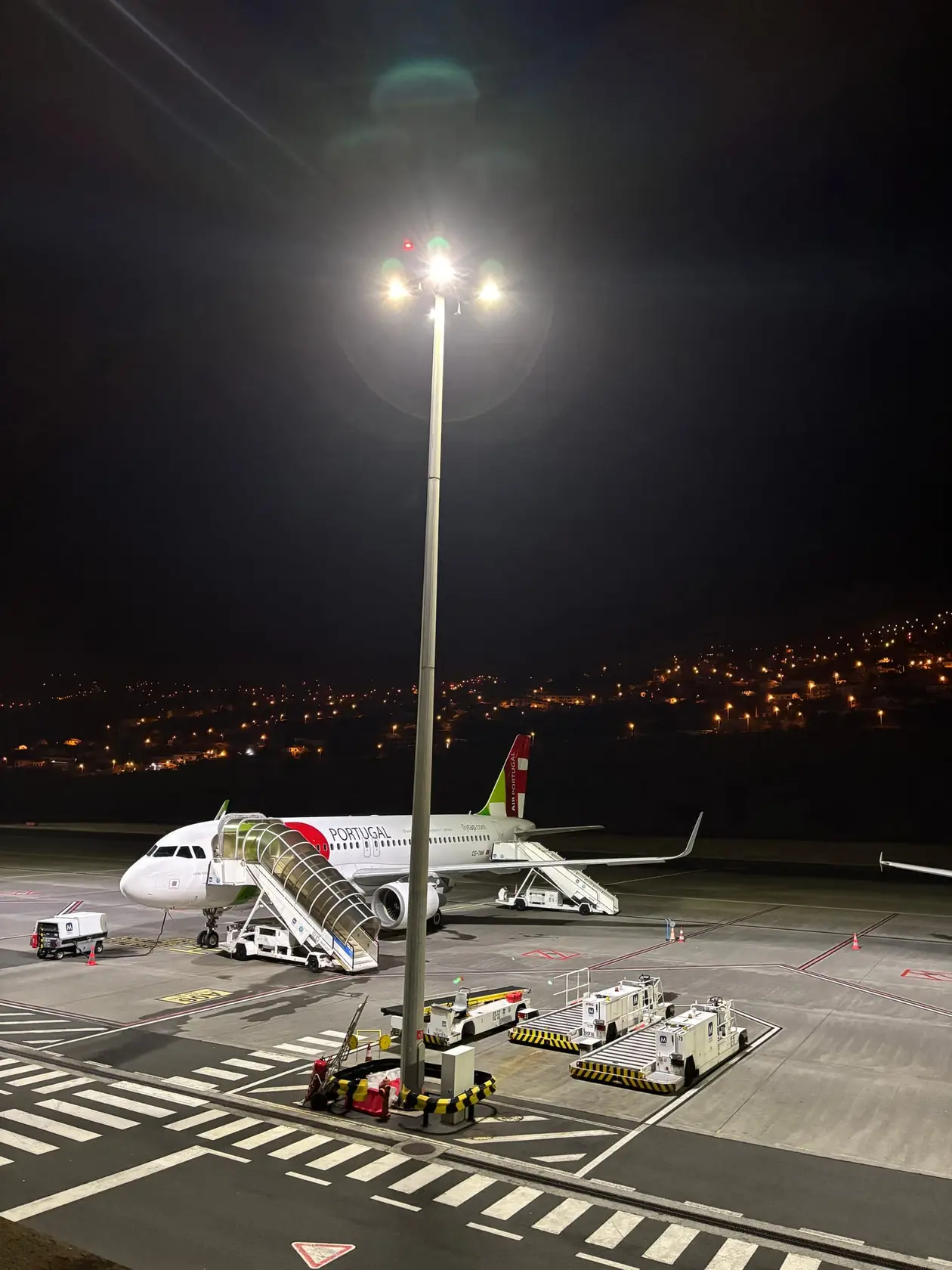 Nachts beleuchtetes Flugzeug mit Gangway auf dem Rollfeld, im Hintergrund die Lichter der Küste