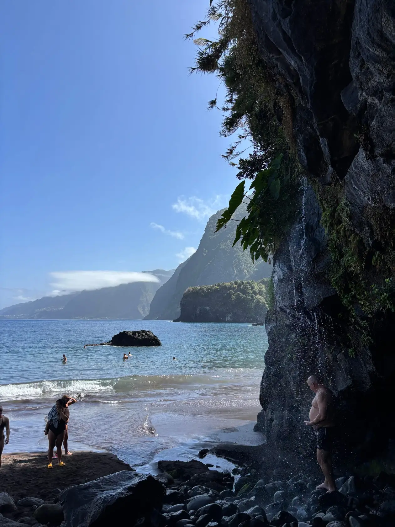 Blick durch eine natürliche Felsöffnung auf eine Bucht mit schwarzem Sand, in der mehrere Personen im Wasser stehen