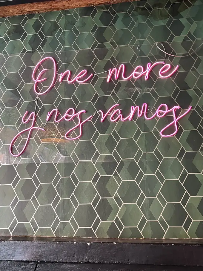 Rosa Neonschrift „One more y nos vamos“ an einer Wand aus grünen Sechseckfliesen