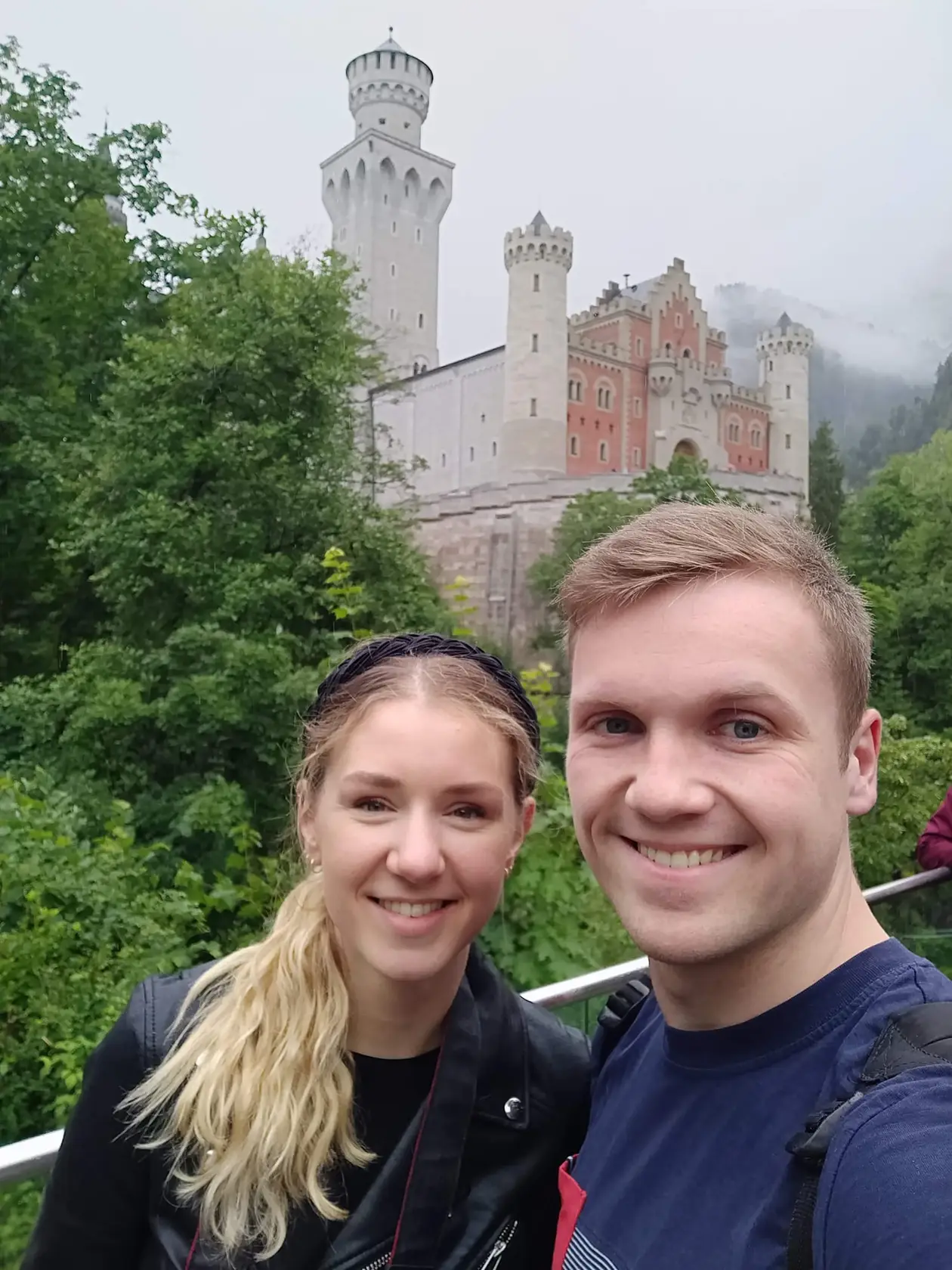 Selfie von Rebecca und Max an einem Geländer, hinter ihnen über den Bäumen Schloss Neuschwanstein mit hellen Türmen und roter Fassade, der Hang dahinter im Nebel