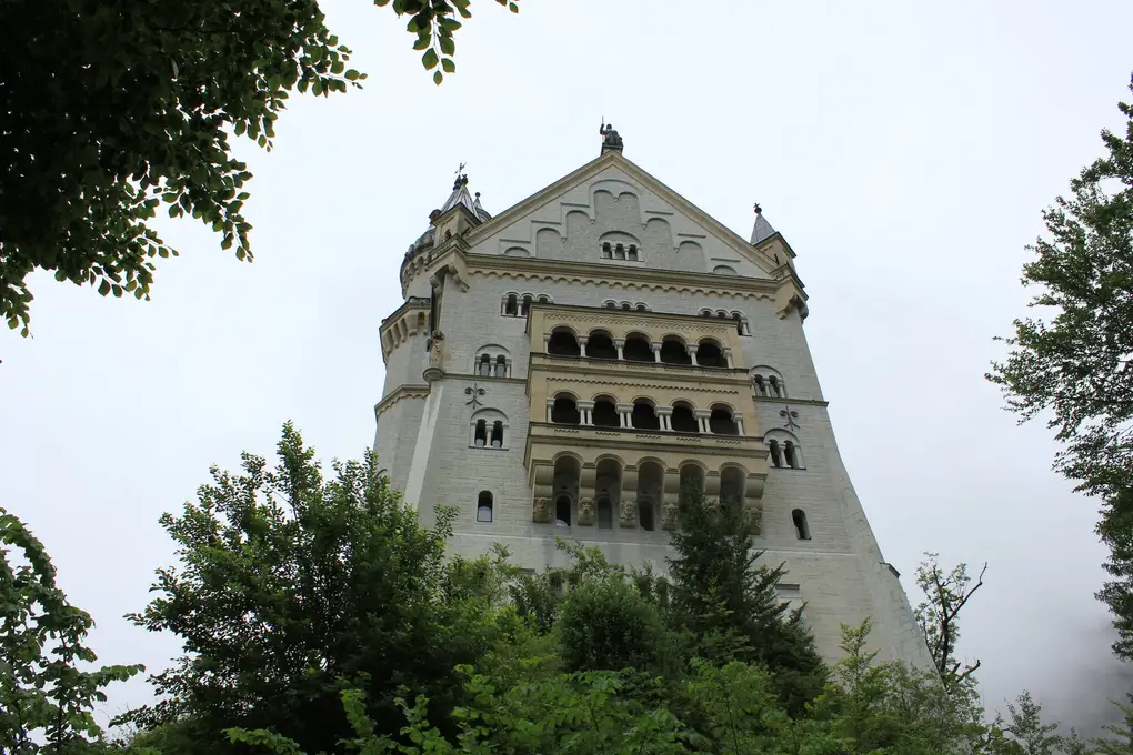 Der hohe Palas von Neuschwanstein steil von unten gesehen, mit Bogenfenstern und Balkonen über mehrere Stockwerke, am Rand Bäume