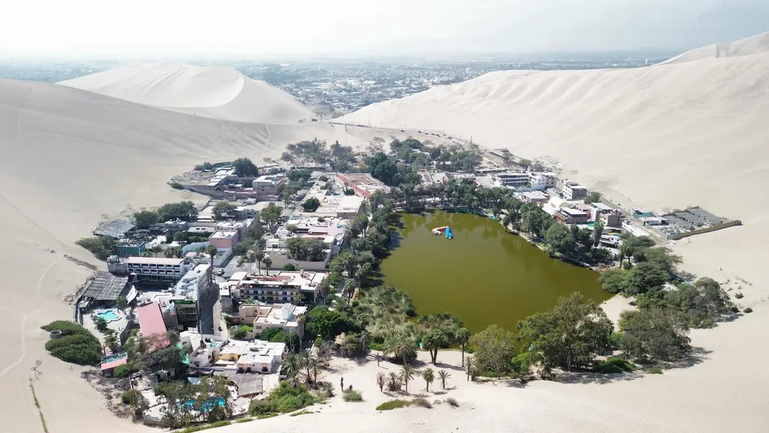 Drohnenbild der Oase Huacachina, eine grüne Lagune mit Palmen und Häusern, ringsum helle Sanddünen und dahinter die Stadt