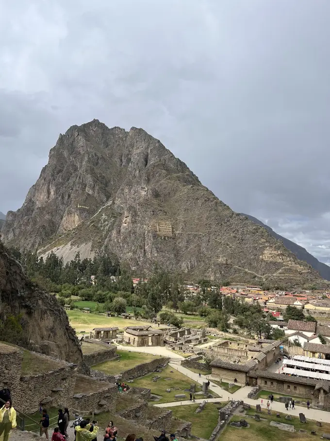 Blick von den Terrassen hinunter auf Ollantaytambo mit Ruinen, Dächern und Bäumen, dahinter ein steiler Felsberg unter grauem Himmel