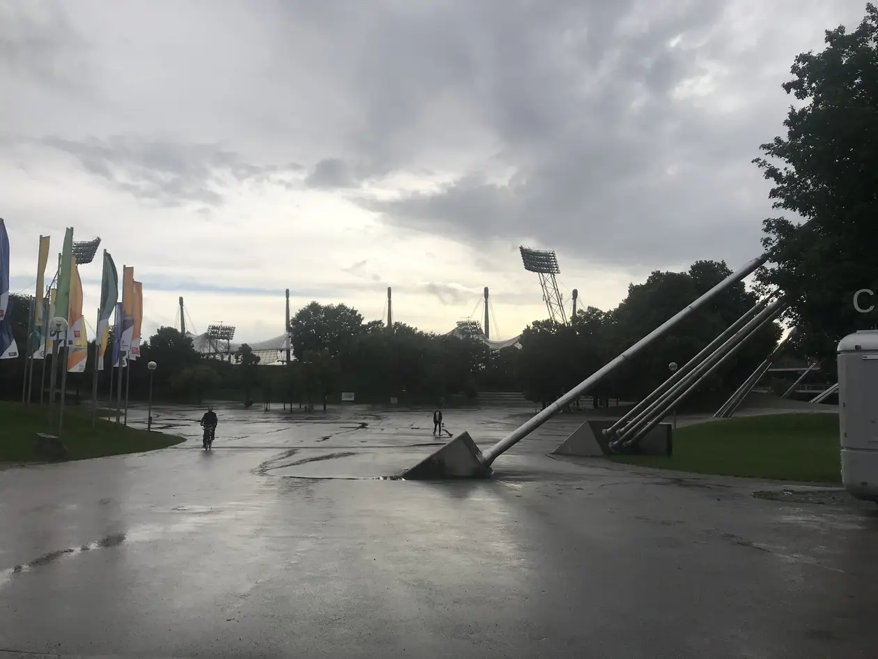 Nasser, weiter Platz im Olympiapark unter grauem Himmel, rechts schräge Stahlstützen, links bunte Fahnen, dahinter über den Bäumen die Masten und Flutlichter des Stadions