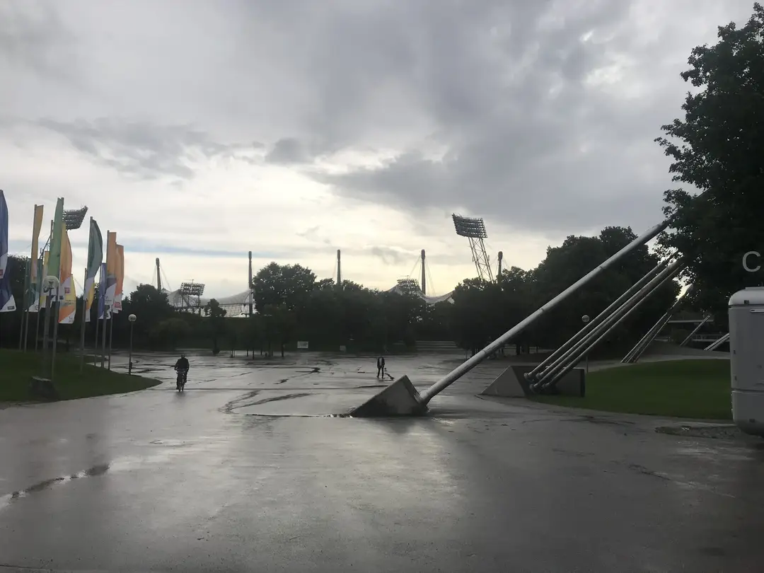 Nasser, weiter Platz im Olympiapark unter grauem Himmel, rechts schräge Stahlstützen, links bunte Fahnen, dahinter über den Bäumen die Masten und Flutlichter des Stadions