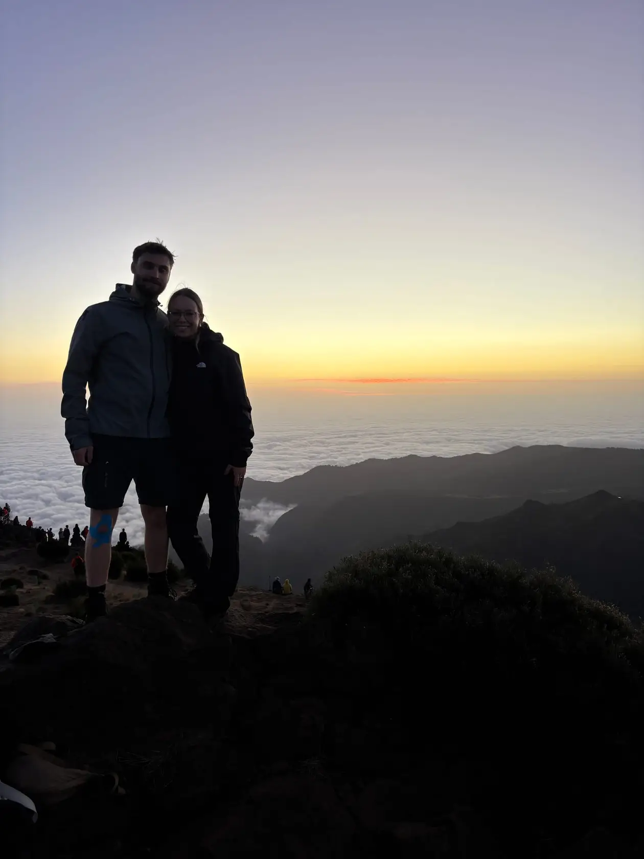 Max und Rebecca stehen als Silhouetten auf einem Felsen über dem Wolkenmeer im Morgenlicht