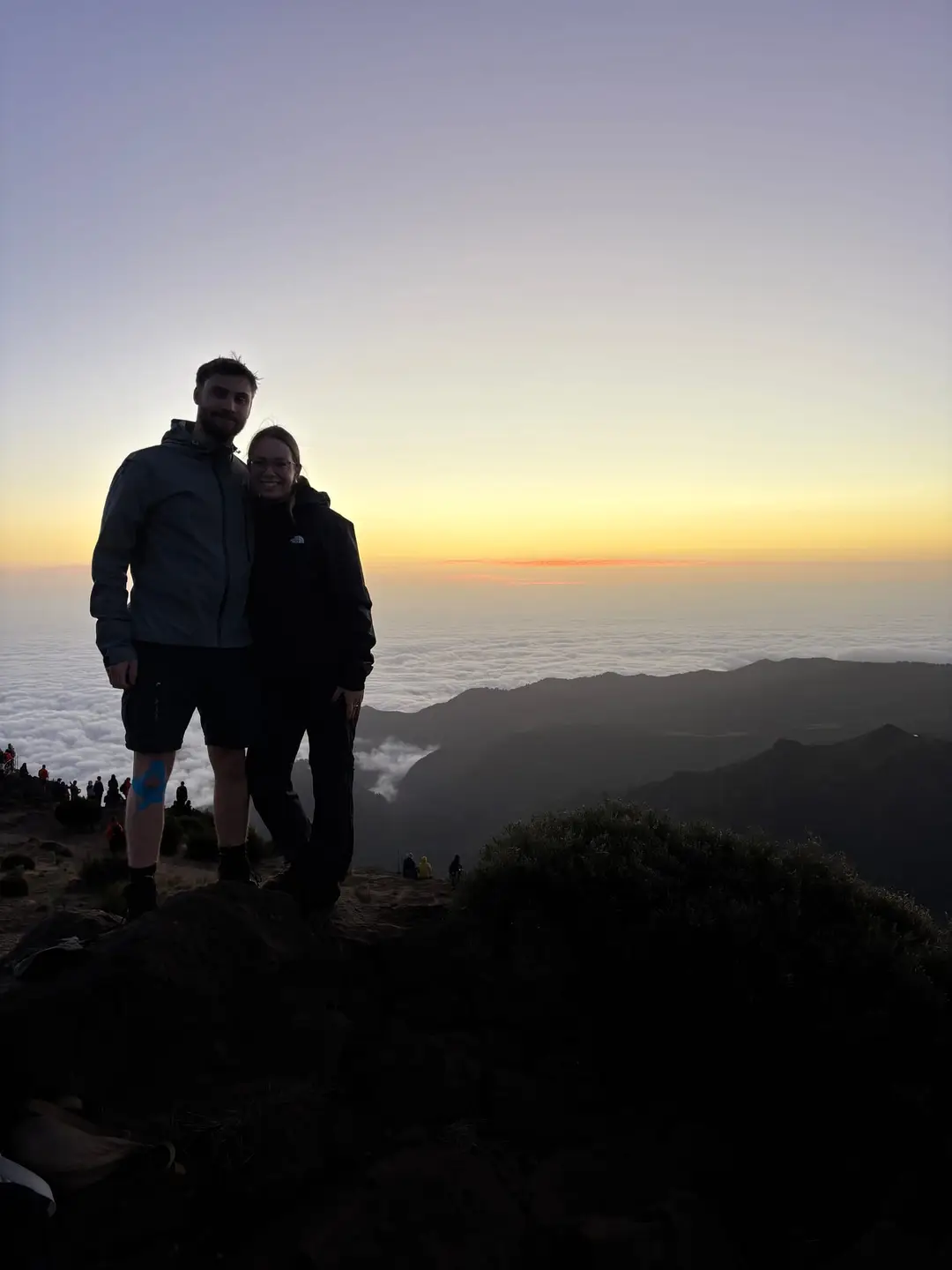 Max und Rebecca stehen als Silhouetten auf einem Felsen über dem Wolkenmeer im Morgenlicht