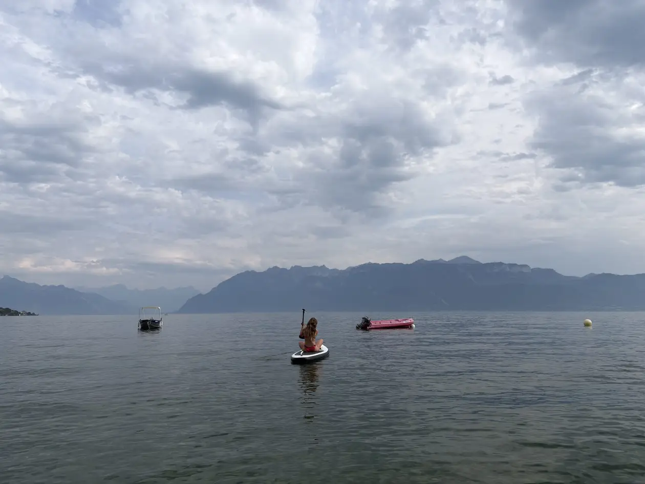 Eine Person kniet auf einem Stand-up-Paddle-Board auf dem Genfersee, daneben Boote, dahinter Berge unter einem bewölkten Himmel