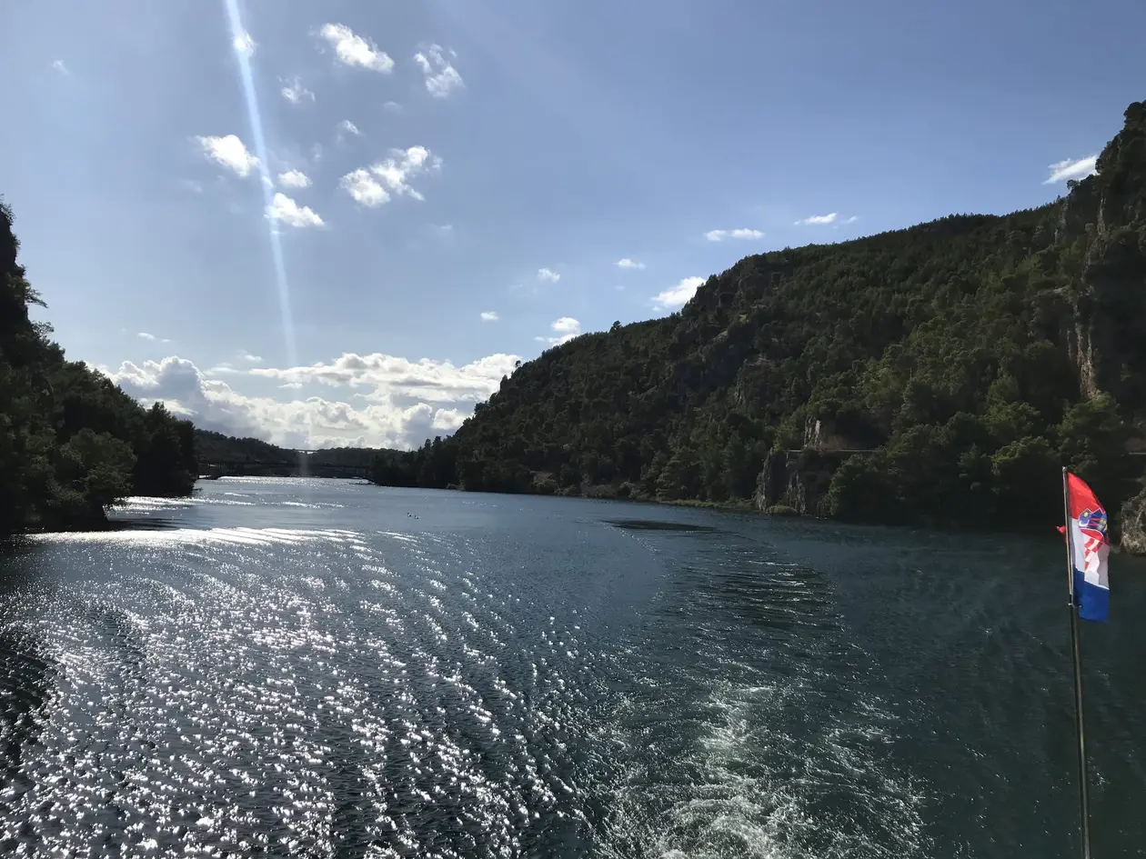 Blick vom Heck eines Boots über das Kielwasser auf den Fluss zwischen felsigen Hängen, am Heck eine kroatische Flagge, hinten eine Brücke