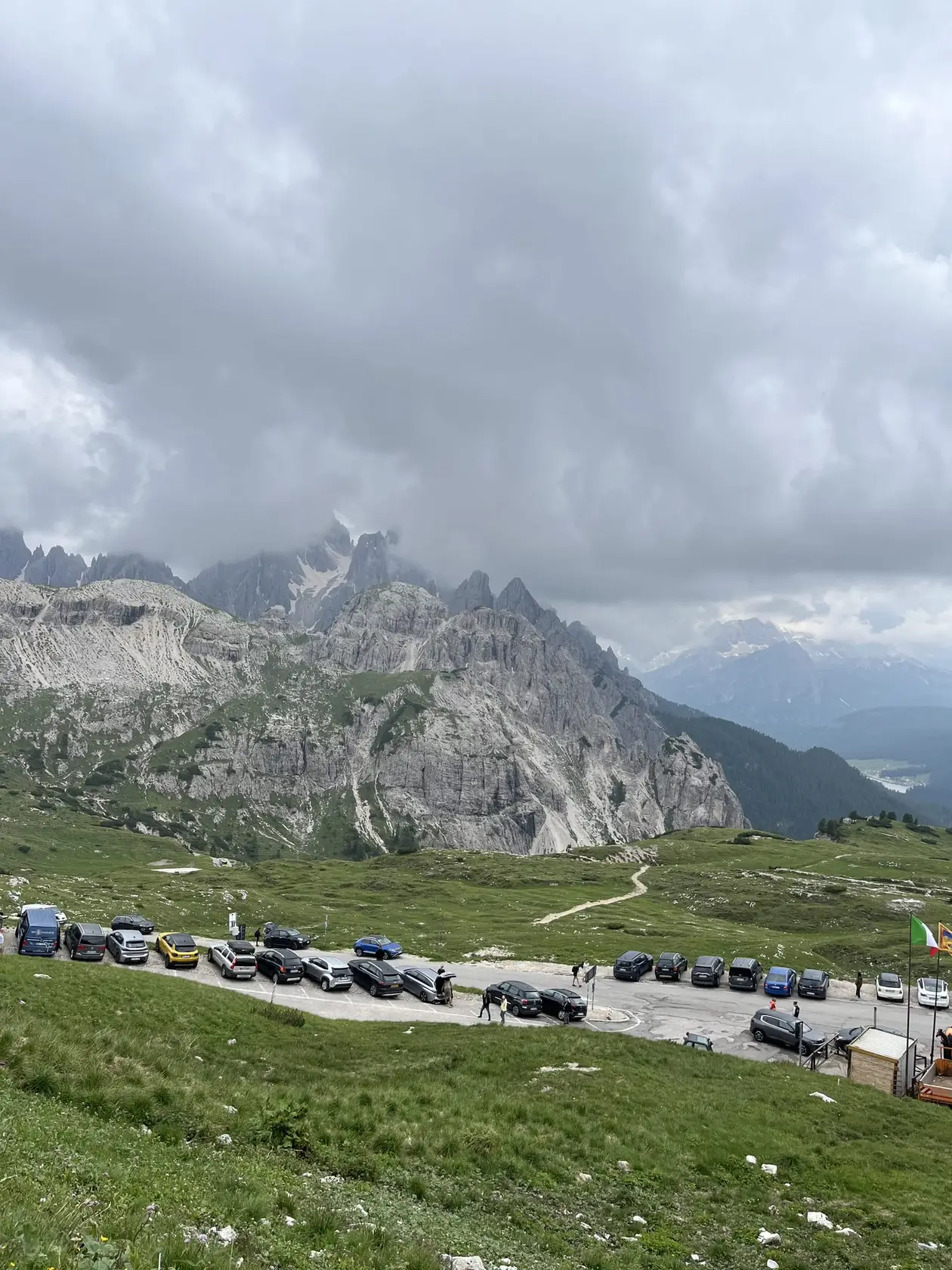 Voller Parkplatz an einer Straßenkehre auf einer Almwiese, dahinter ein Wanderweg und zerklüftete Berge in grauen Wolken, rechts unten im Tal ein See