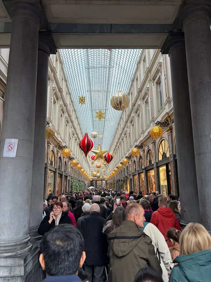 Blick zwischen zwei Säulen in eine überfüllte Einkaufspassage mit Glasdach, darunter hängen große rote und goldene Weihnachtskugeln und Sterne