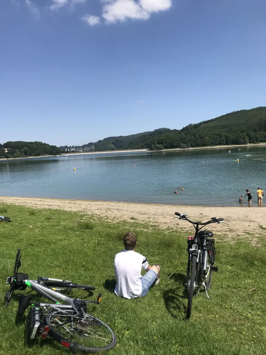 Max sitzt von hinten gesehen im Gras am Ufer des Hennesees, links liegt ein E-Bike mit Akku am Gepäckträger, rechts steht ein zweites, vorne ein Kiesstrand und Badende im See