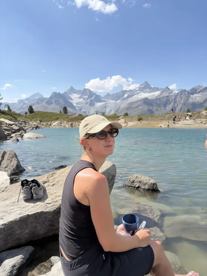 Rebecca sitzt mit Kappe auf einem Felsen am Ufer eines türkisen Bergsees, neben ihr ein Paar Schuhe, dahinter verschneite Berge