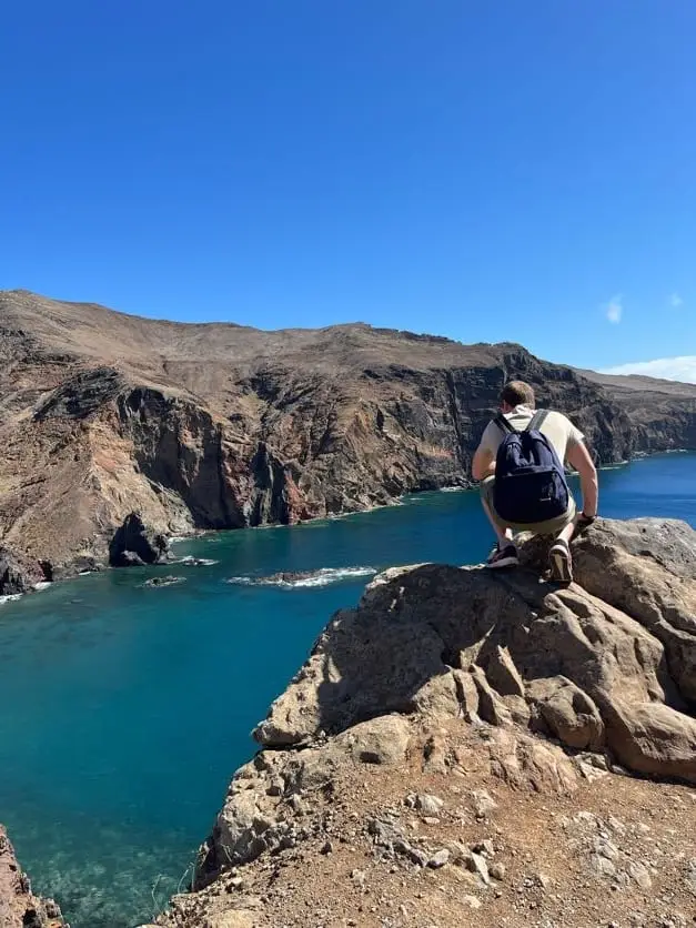 Ein Wanderer mit Rucksack sitzt auf einem Felsen über einer türkisfarbenen Bucht