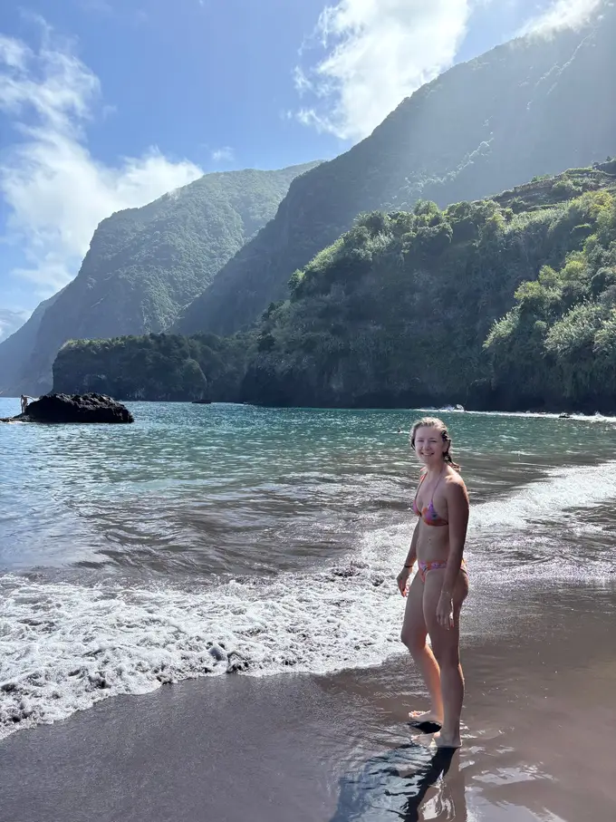 Eine Person im Bikini steht in der Brandung am schwarzen Sandstrand, dahinter steile bewaldete Klippen