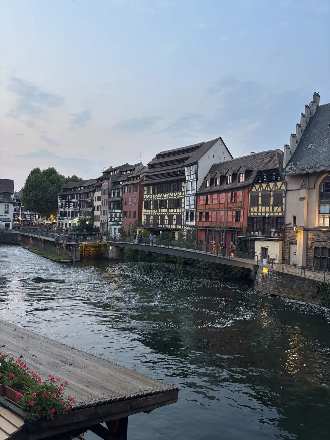 Bunte Fachwerkhäuser am Ufer der Ill in der Abenddämmerung, davor ein Steg und eine Fußgängerbrücke mit Spaziergängern