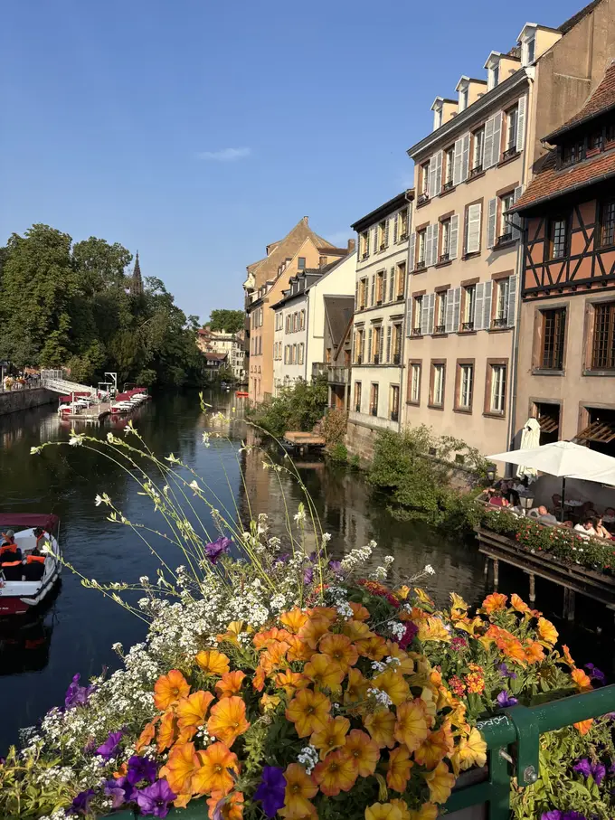 Blick von einer Brücke mit orangefarbenen Blumen auf einen Kanal, rechts Häuser mit Fensterläden und eine Restaurantterrasse über dem Wasser, links Boote