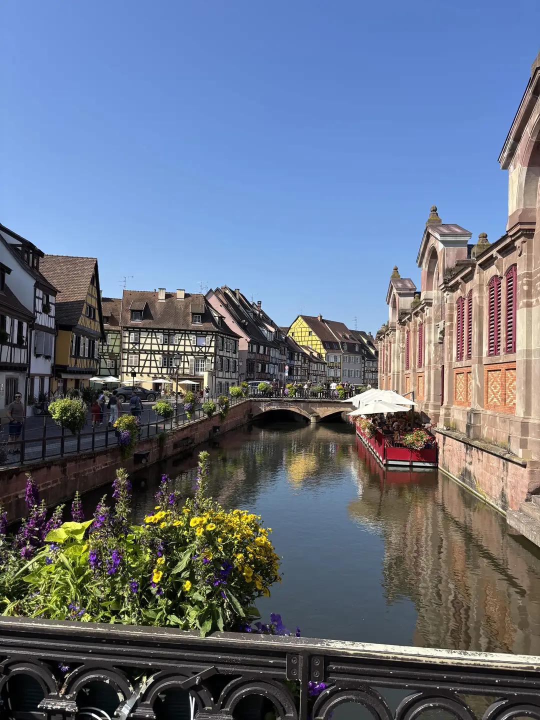 Blick von einer Brücke mit gelben und lila Blumen auf einen Kanal zwischen bunten Fachwerkhäusern, rechts ein Gebäude aus rotem Sandstein, davor eine Restaurantterrasse am Wasser