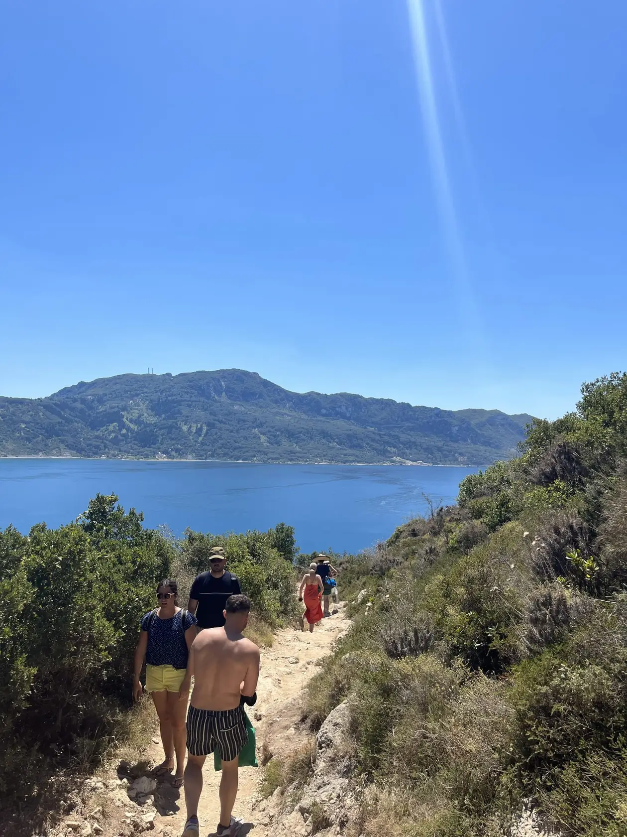 Schmaler, steiniger Pfad zwischen Büschen, darauf Wanderer, unten eine Bucht und dahinter Berge